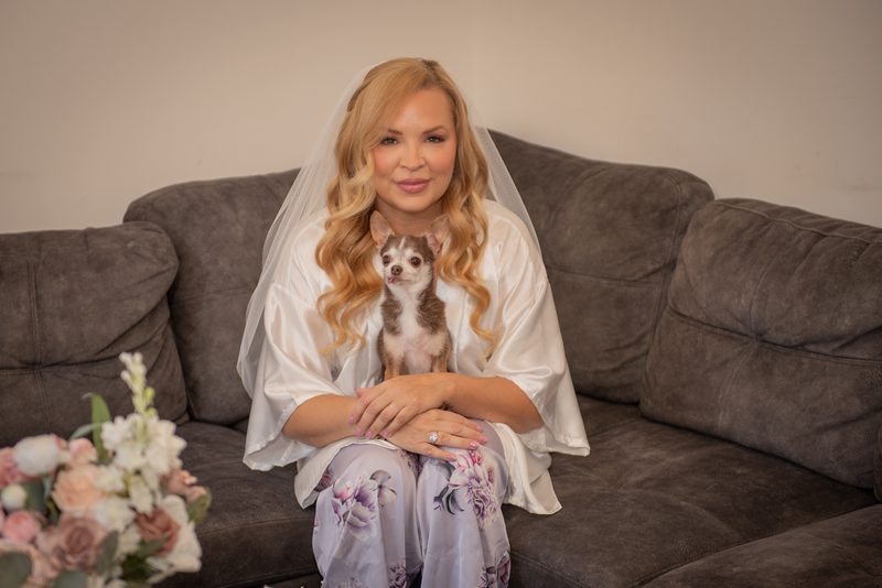 A bride in a white satin robe and veil sits on a sofa with a chihuahua, smiling gently at the camera.