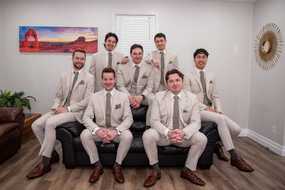 Seven men in beige suits and gray ties pose formally around a black leather couch in a warmly lit room, smiling.