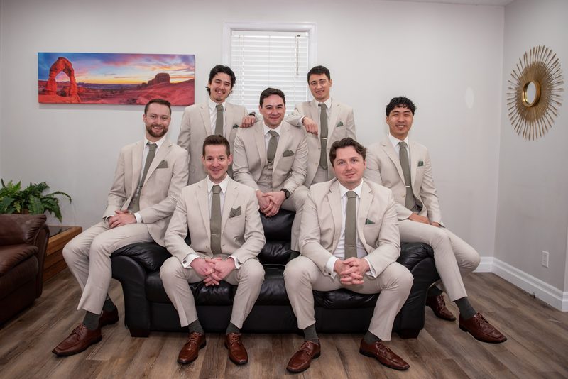 Seven men in beige suits and gray ties pose formally around a black leather couch in a warmly lit room, smiling.