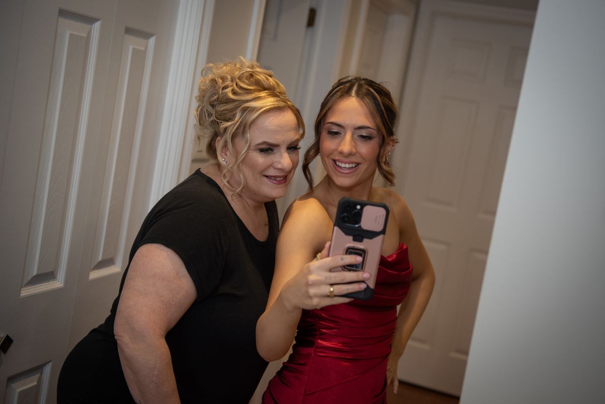 Two women, one in a red satin gown and the other in a black top, smile for a selfie in a hallway during a celebration.