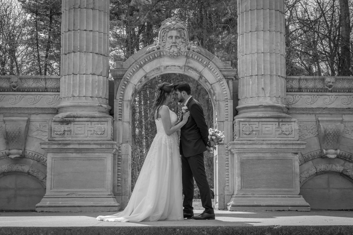 A bride and groom stand forehead to forehead beneath an ornate stone archway, in black and white with bare trees behind them.