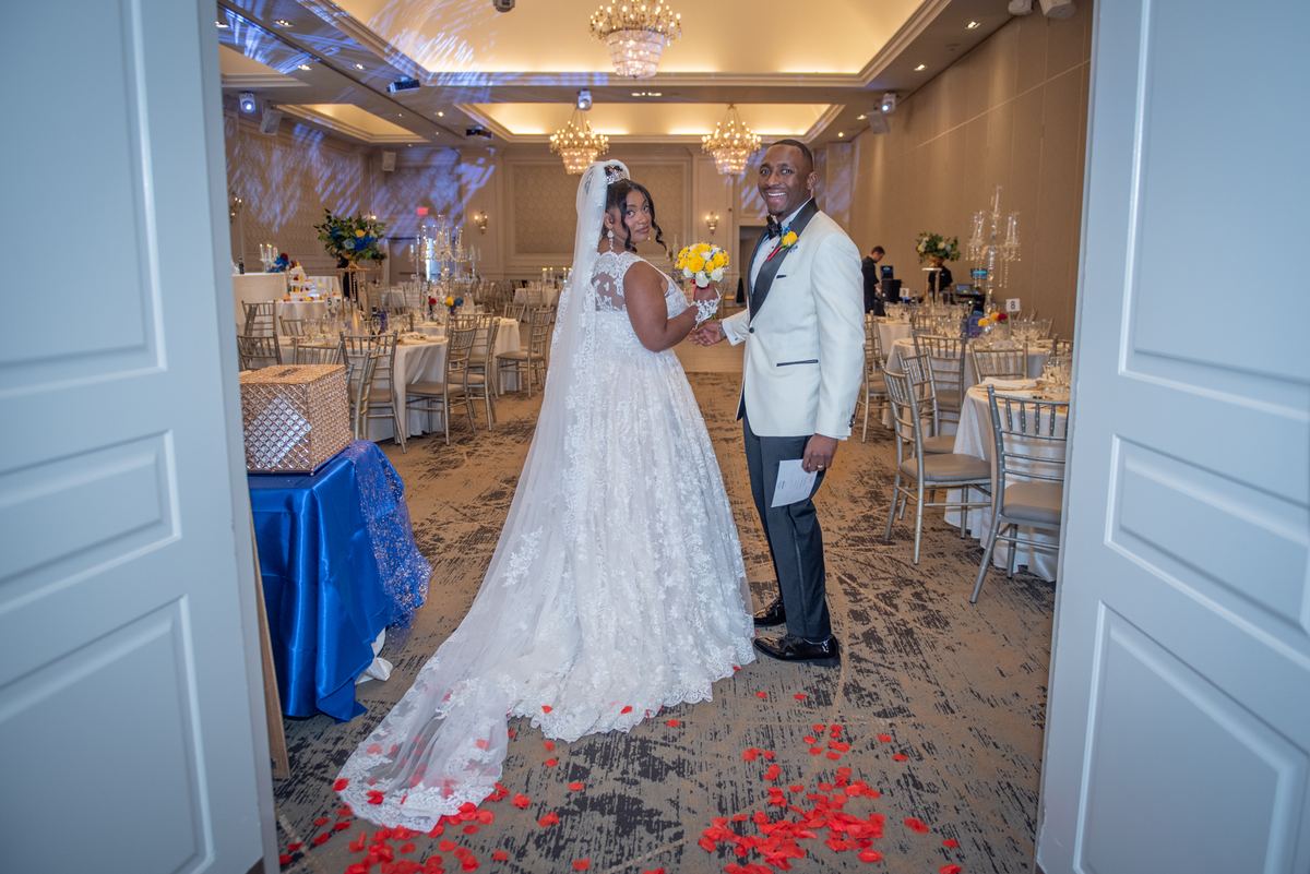 Bride and groom smile as they stand in a grand hall with rose petals at their feet, holding bouquet and program.