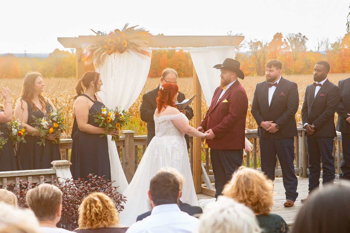 A couple exchanges vows under a wooden arch adorned with autumn flowers and white drapery, surrounded by fall foliage.