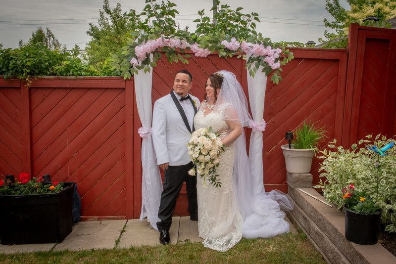 A smiling bride and groom stand under a decorated arch in a backyard garden, framed by red fencing and potted plants.