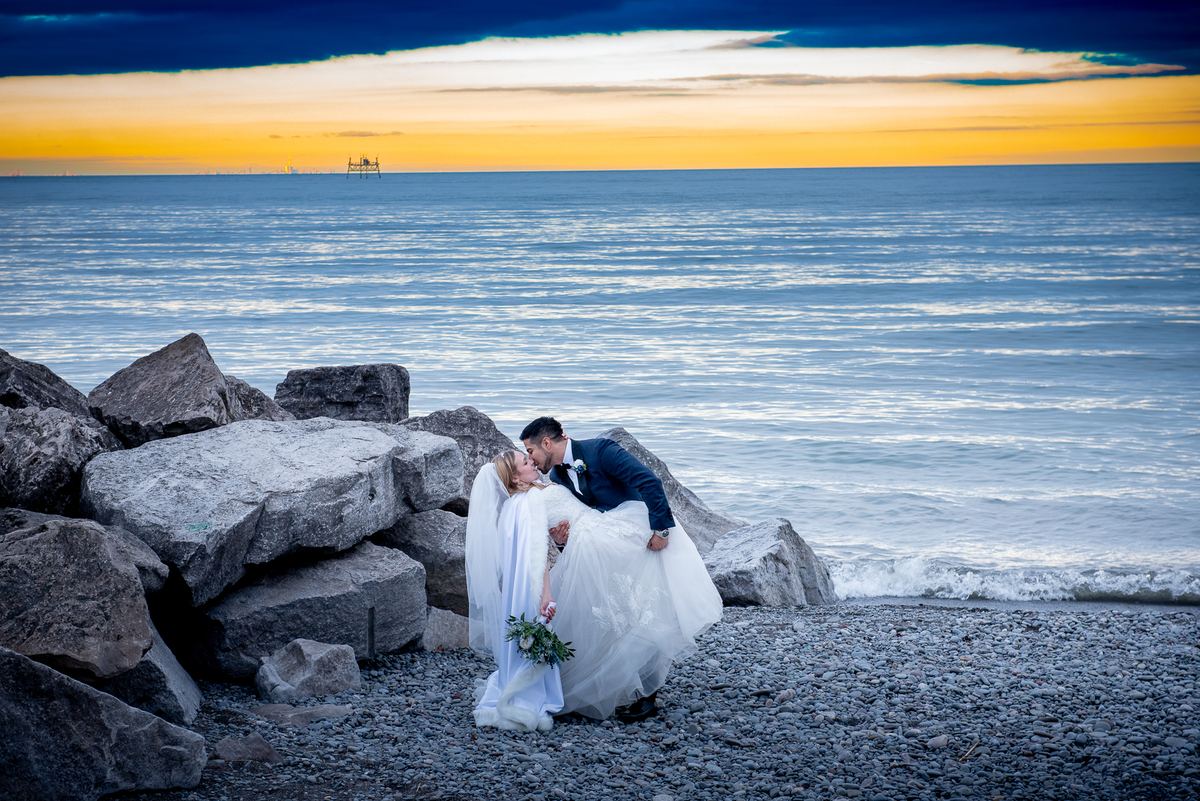 A newlywed couple kisses on a rocky shoreline as the sun sets, casting golden light over the water and sky.