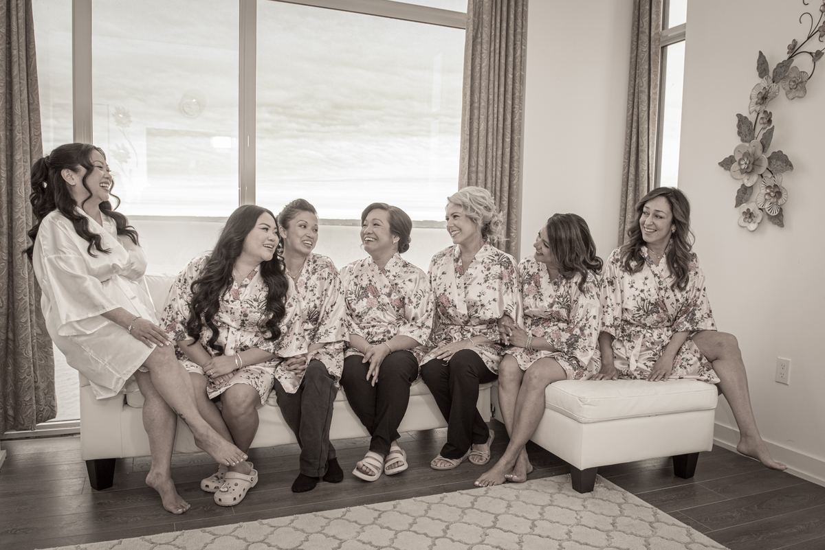 A bride and her bridesmaids laugh together on a white sofa in a modern room filled with natural light.