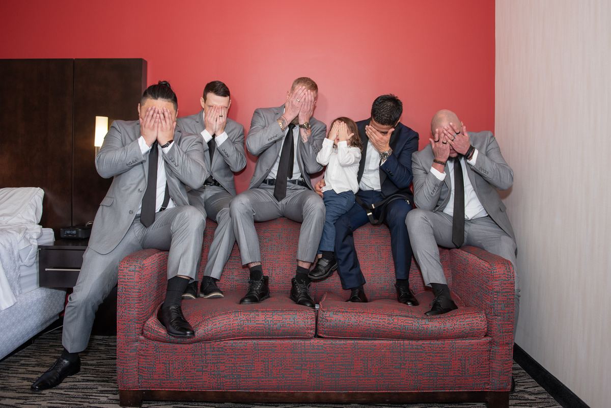 Six formally dressed people, including one girl, sit on a red patterned couch with eyes covered, creating a playful moment.