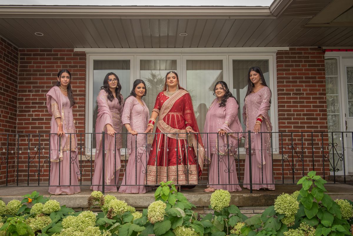 A bride in red and gold stands with five bridesmaids in dusty pink on a porch, framed by green hydrangeas.