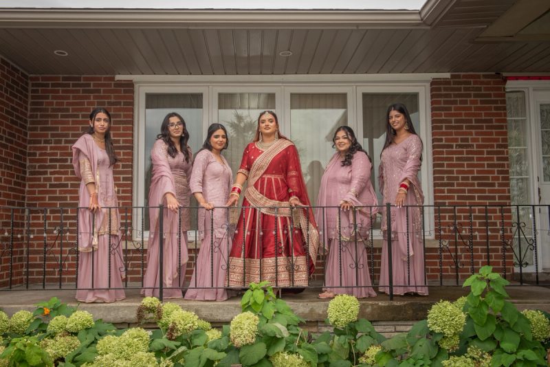 A bride in red and gold stands with five bridesmaids in dusty pink on a porch, framed by green hydrangeas.