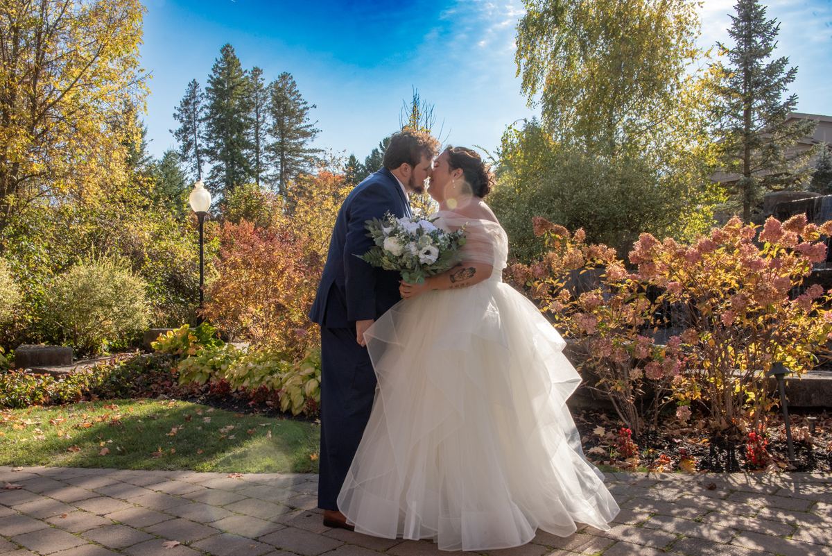A newlywed couple exchanges kisses outdoors in autumn, surrounded by colorful leaves and a clear sky.