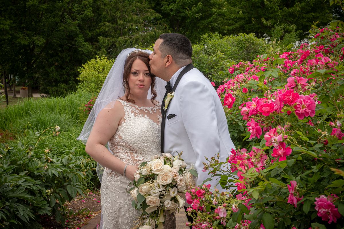 A groom in a white tuxedo kisses the bride's cheek as she holds roses and smiles at the camera, framed by greenery.