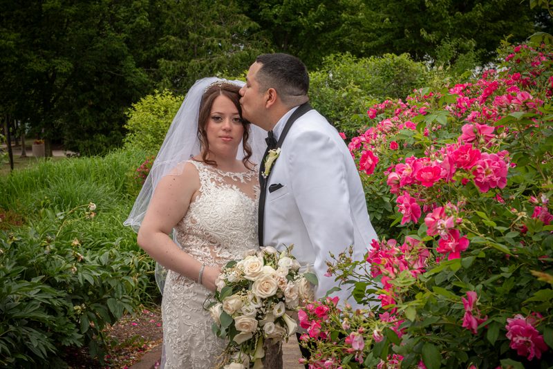 A groom in a white tuxedo kisses the bride's cheek as she holds roses and smiles at the camera, framed by greenery.