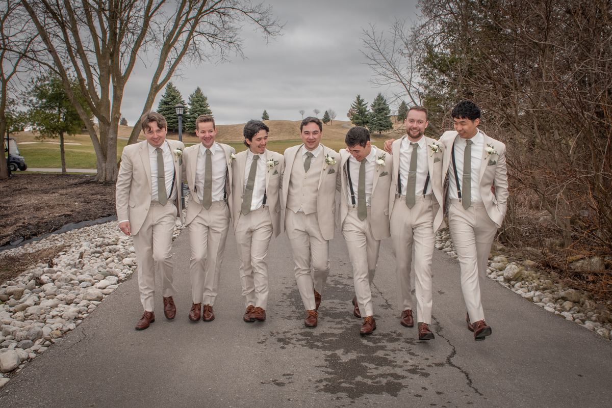 Seven groomsmen in beige suits and brown shoes walk along a paved path, laughing under bare trees on a cool day.