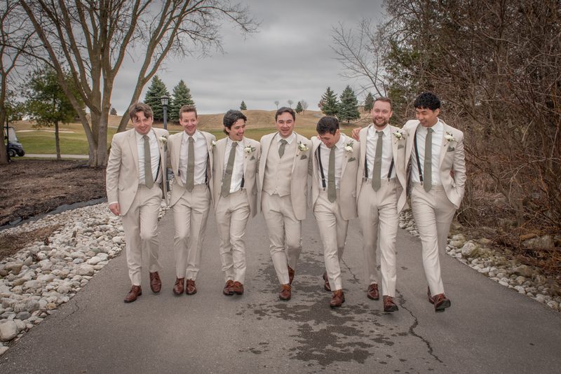 Seven groomsmen in beige suits and brown shoes walk along a paved path, laughing under bare trees on a cool day.