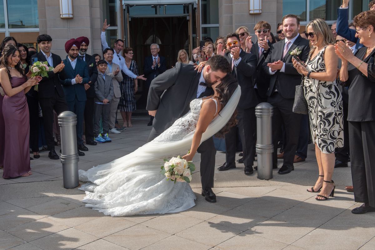 A newlywed couple kisses outside a building as smiling guests in formal attire clap and take photos, with the bride's veil.