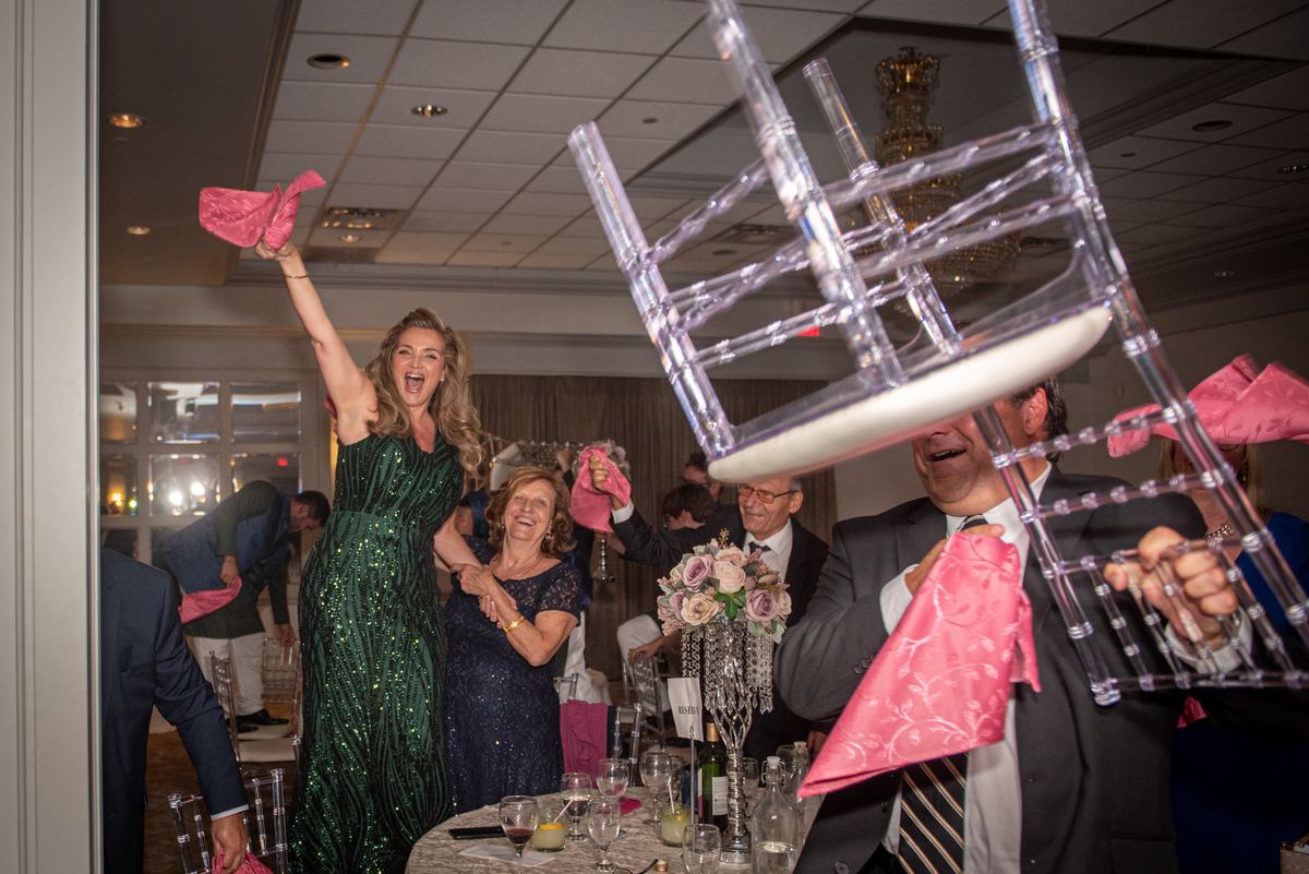 A woman in a green sequined gown and a man in a suit playfully hold pink napkins and an overturned chair as guests laugh.