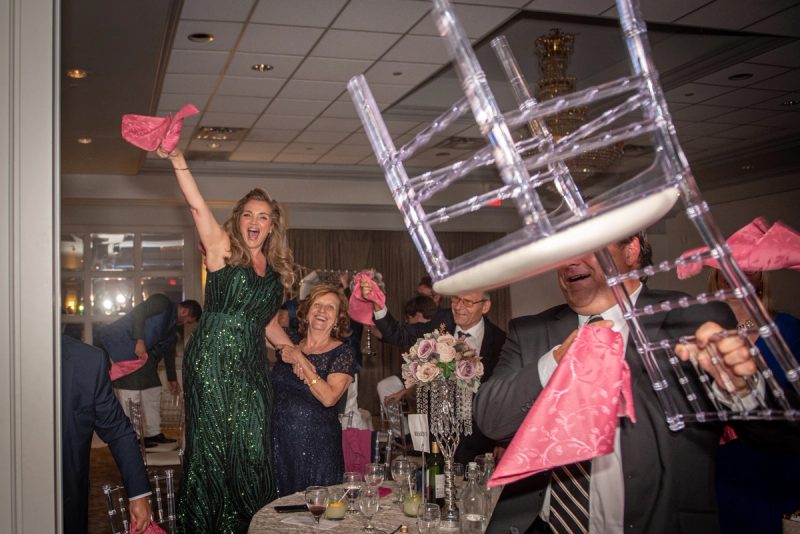 A woman in a green sequined gown and a man in a suit playfully hold pink napkins and an overturned chair as guests laugh.