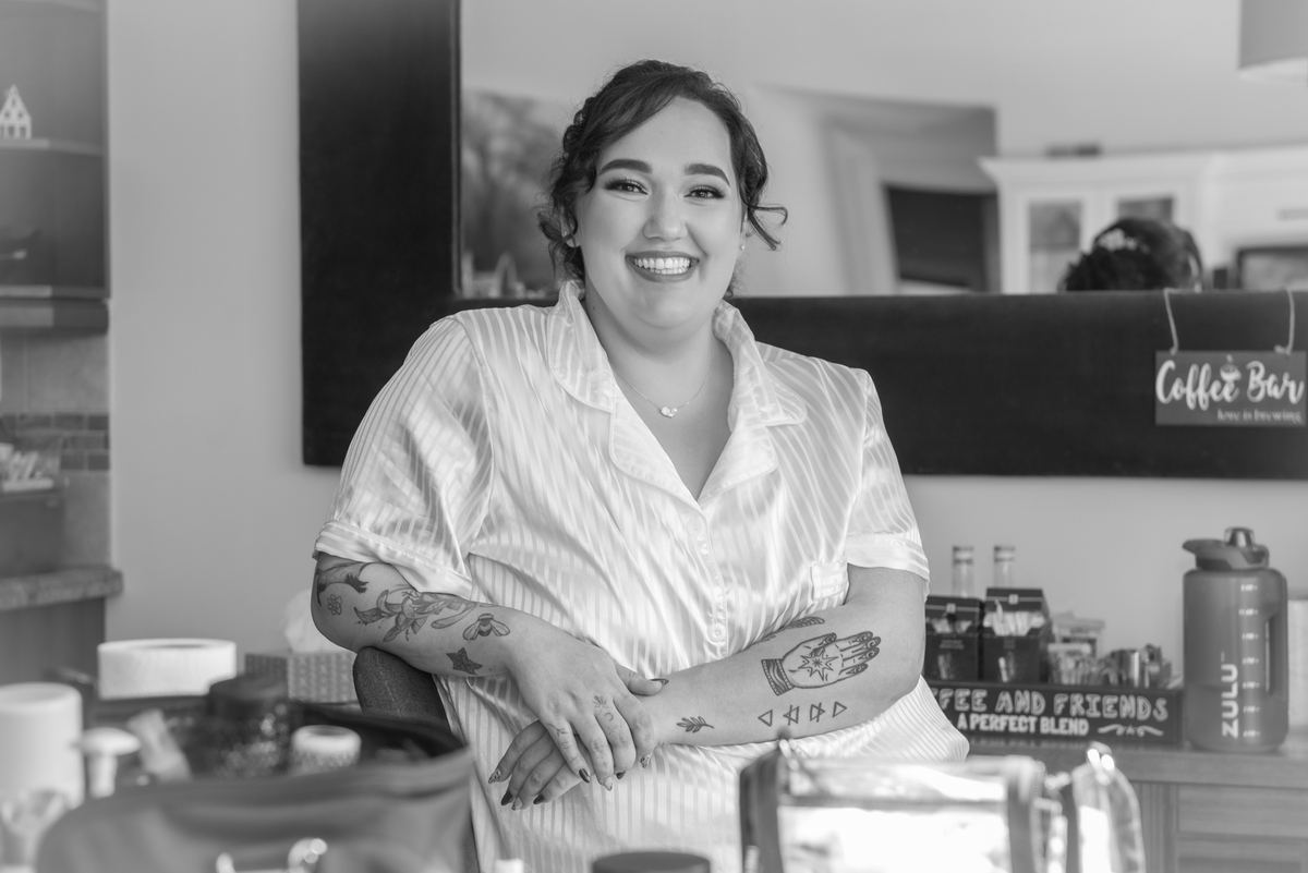 A smiling woman with tattoos stands behind a coffee bar in a cozy café with soft lighting and cluttered shelves.