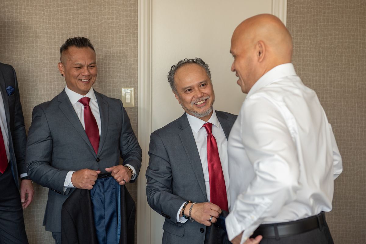 Three formally dressed men in red ties laugh together, one adjusting his jacket, creating a joyful pre-wedding atmosphere.