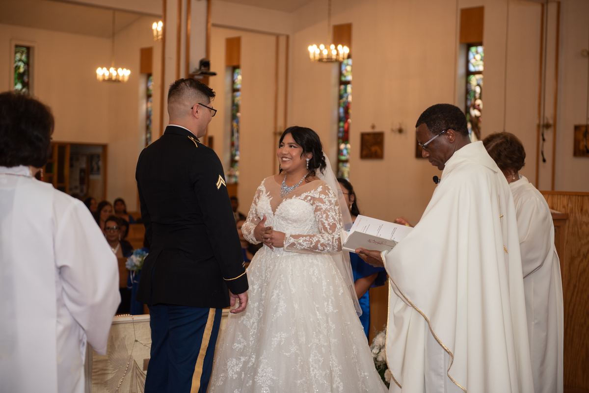 A smiling bride in a lace gown stands beside her groom in military uniform during a church ceremony.