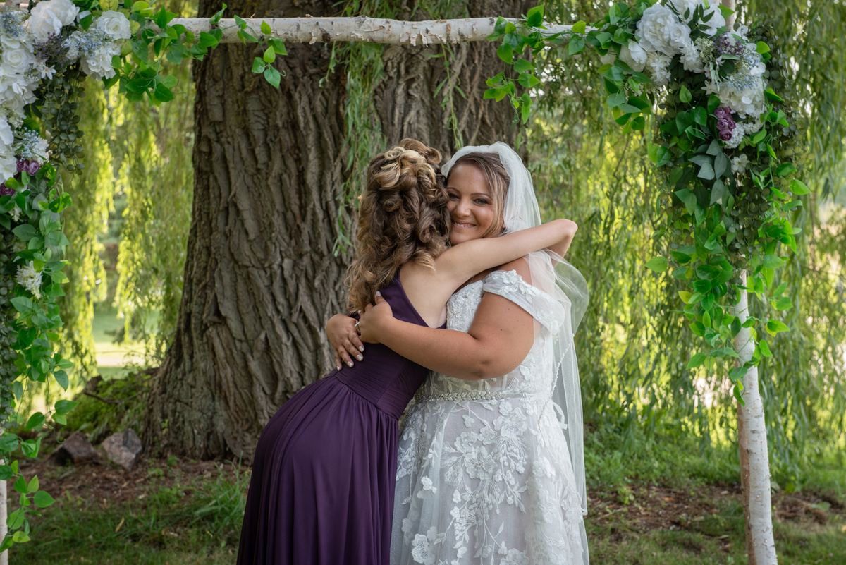A bride in white lace embraces a bridesmaid in purple under a floral arch, sunlight filtering through trees.