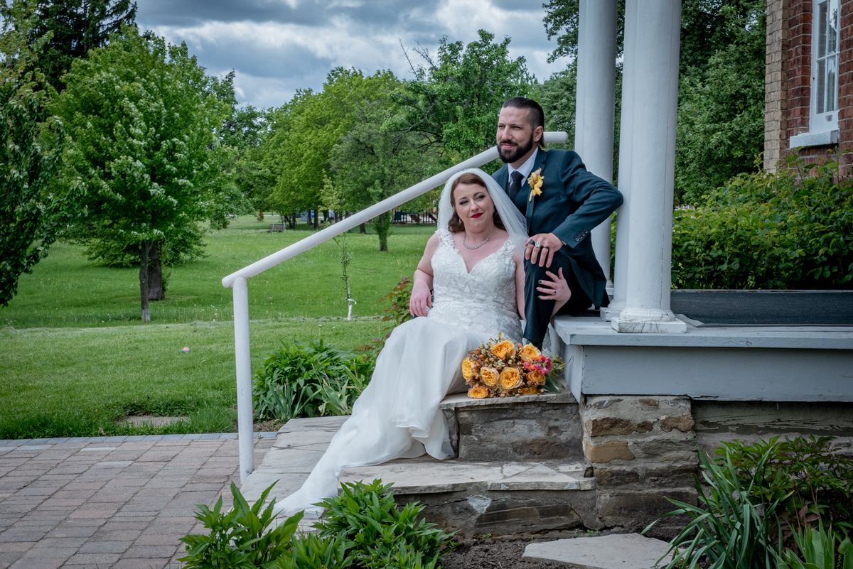 Bride and groom sit relaxed on stone steps surrounded by lush greenery under an overcast sky, bouquet in hand.
