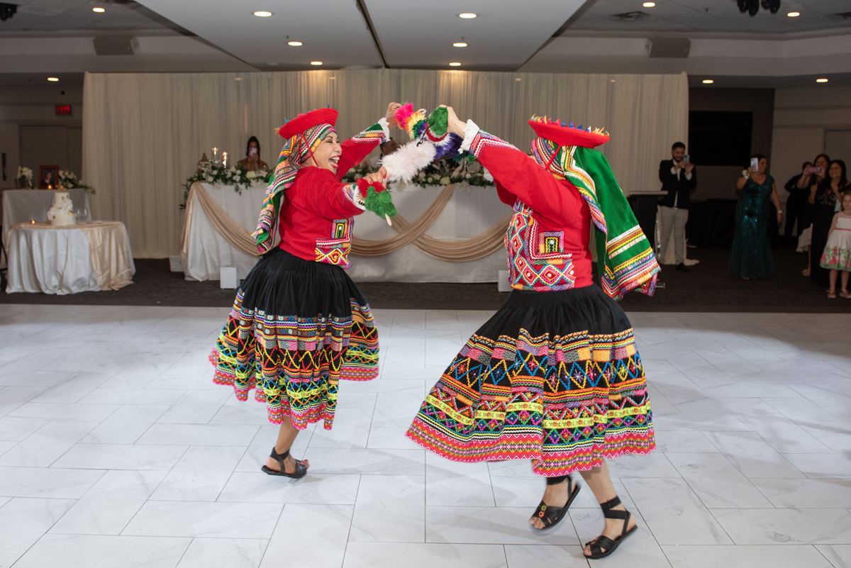 Two women in traditional Andean attire dance joyfully, exchanging feathered accessories on a ballroom floor.