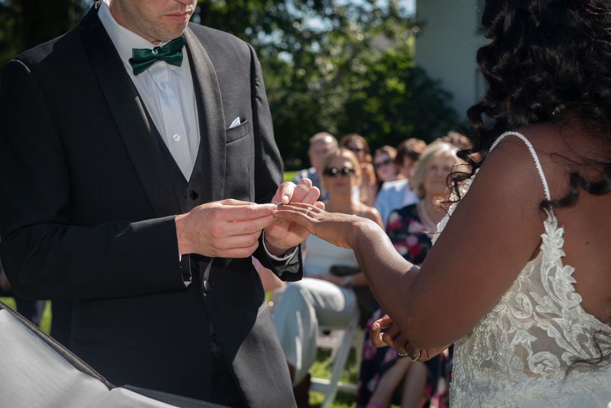 Groom in black tuxedo with green bow tie places ring on bride's outstretched hand, guests blurred in background.