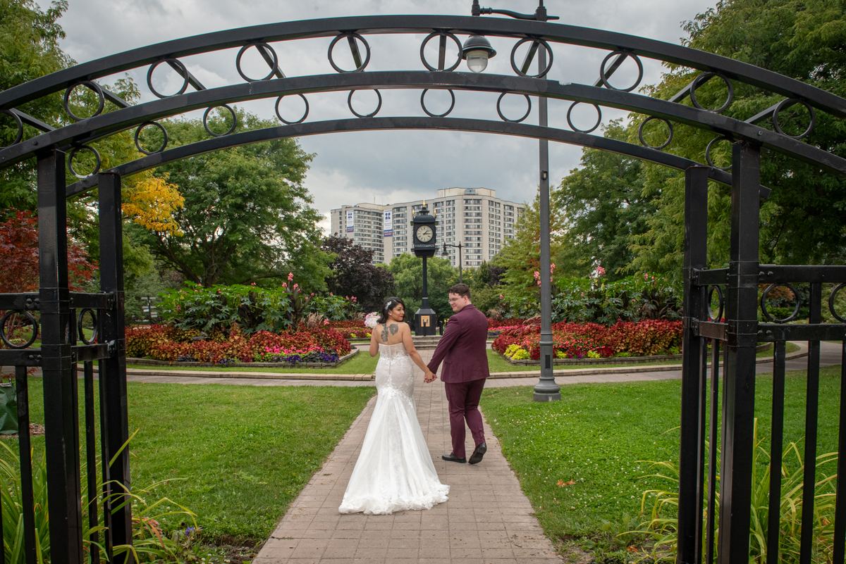 A bride and groom walk hand-in-hand under an ornate black metal arch in a lush park with colorful flowers.