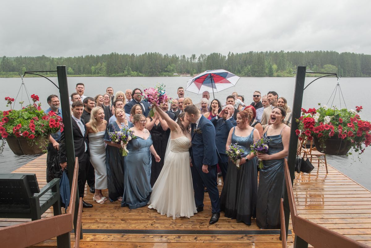 A bride and groom kiss on a wooden dock by a lake, surrounded by guests holding umbrellas in the rain.