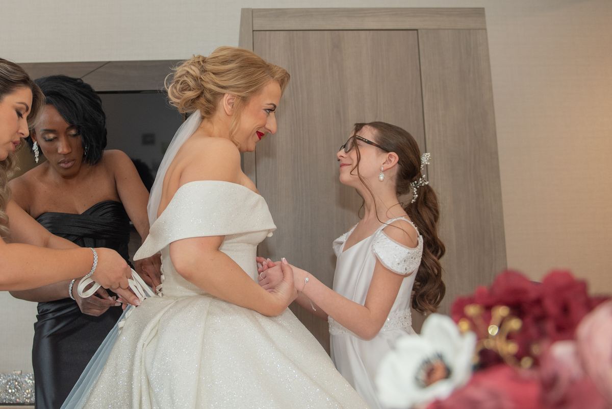 A bride in an off-the-shoulder gown is helped by a woman in black while a young girl holds her hand.
