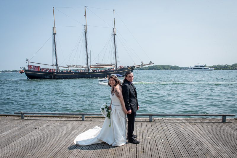 A newlywed couple smiles on a wooden dock with a historic sailing ship and modern yacht in the background, water calm.