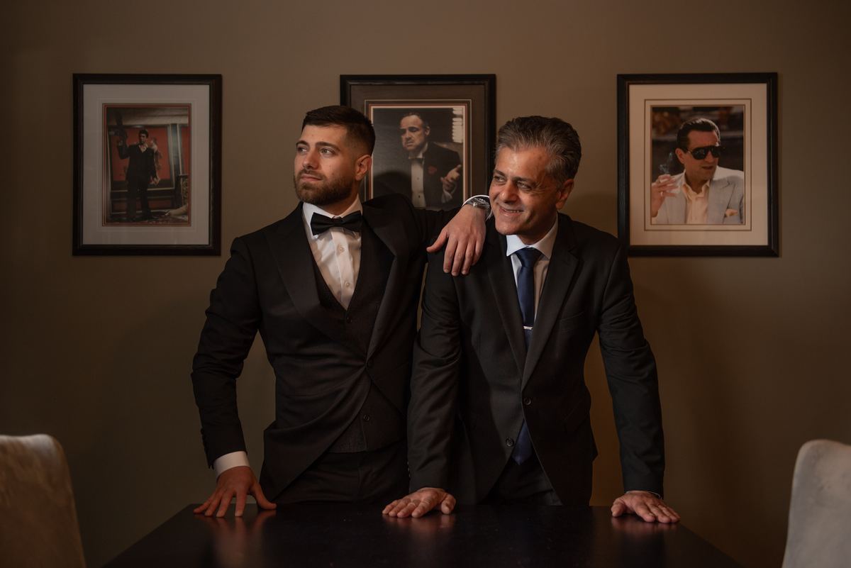 Two formally dressed men pose seriously and smilingly beside a table with gangster-themed wall art behind them.