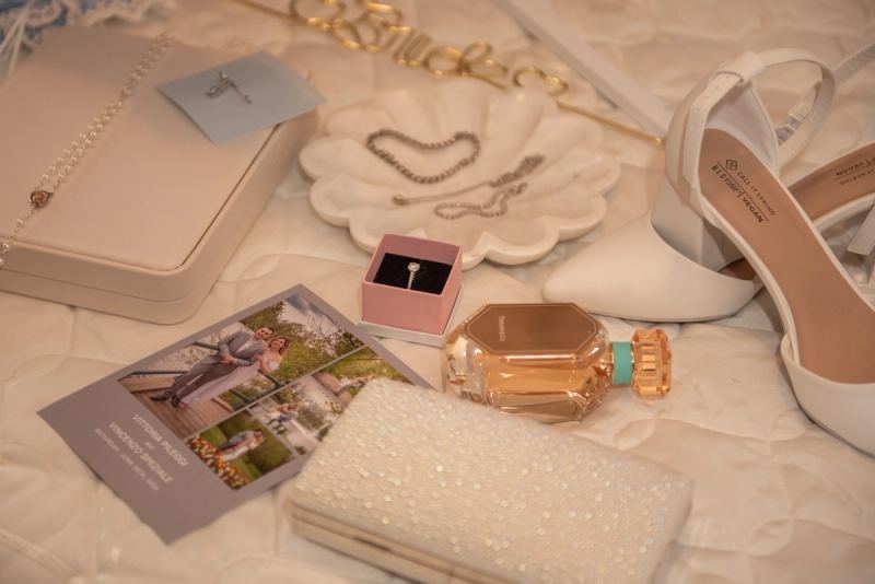 Wedding accessories including a clutch, perfume bottle, heels, and photo card are arranged on a textured surface.