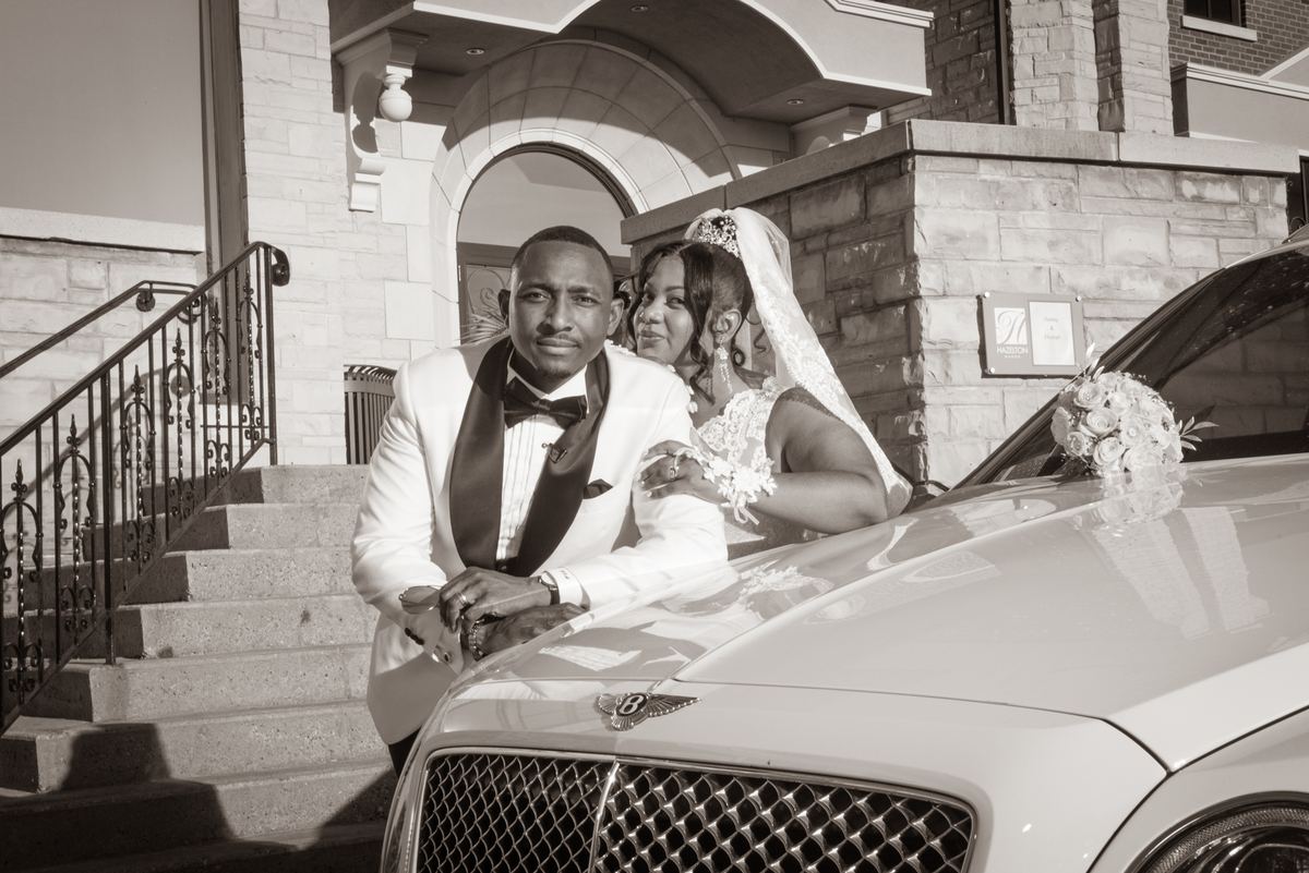 A newlywed couple poses joyfully beside a luxury Bentley, the bride leaning on the hood as sunlight casts sharp shadows.