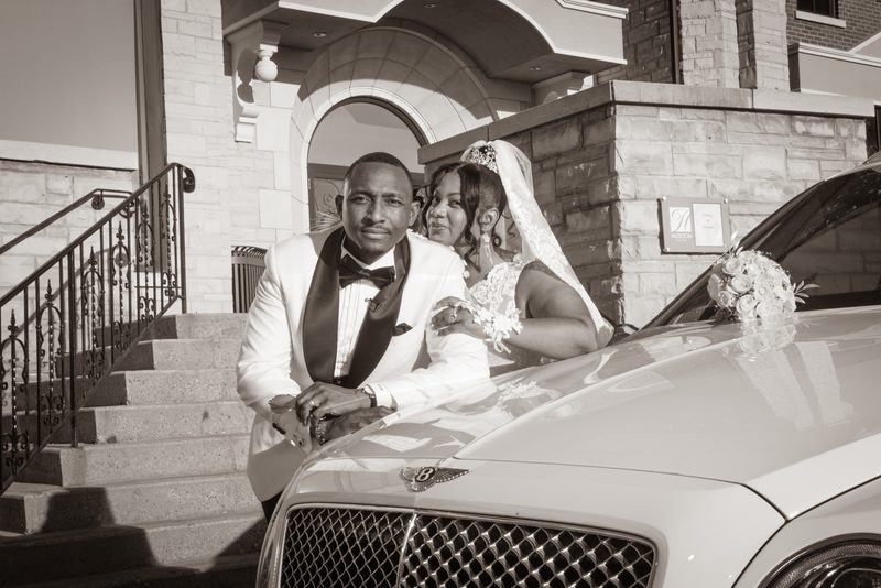 A newlywed couple poses joyfully beside a luxury Bentley, the bride leaning on the hood as sunlight casts sharp shadows.