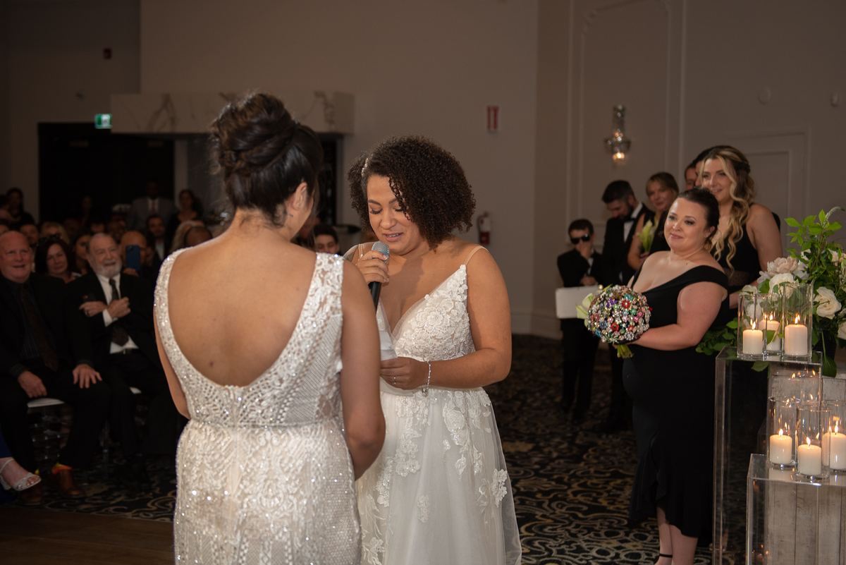 A bride in a sparkling gown listens as her partner speaks with emotion during an indoor ceremony, surrounded by guests.