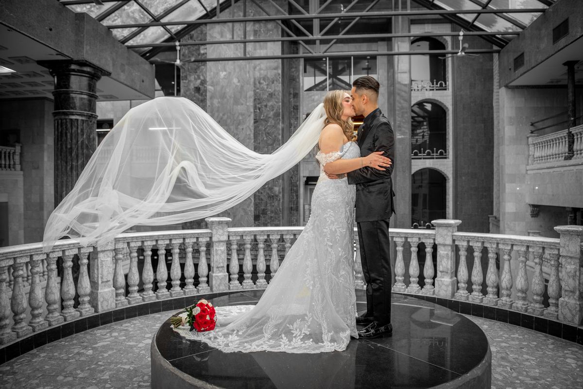 Bride and groom kiss on marble platform in ornate atrium, her veil billowing, with red bouquet at their feet.