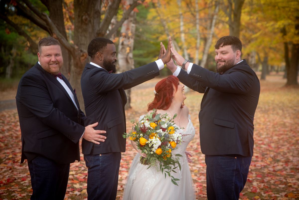 A red-haired bride in a white gown high-fiving three groomsmen amid autumn foliage and fallen leaves.