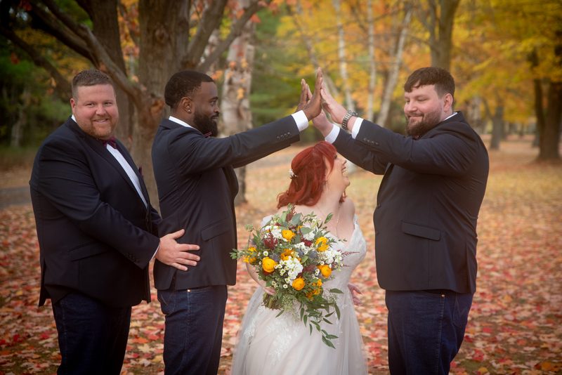 A red-haired bride in a white gown high-fiving three groomsmen amid autumn foliage and fallen leaves.