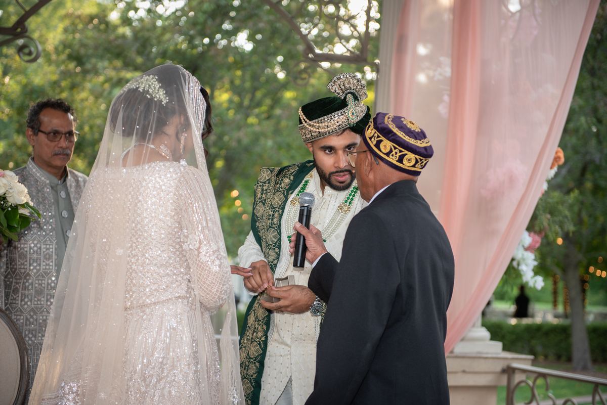 Bride in white and groom in traditional attire exchange rings under dappled sunlight during ceremony.