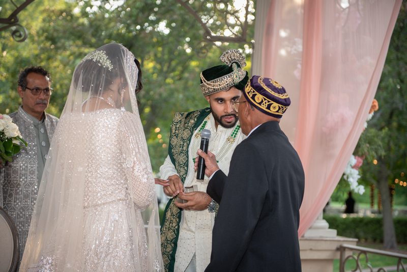 Bride in white and groom in traditional attire exchange rings under dappled sunlight during ceremony.