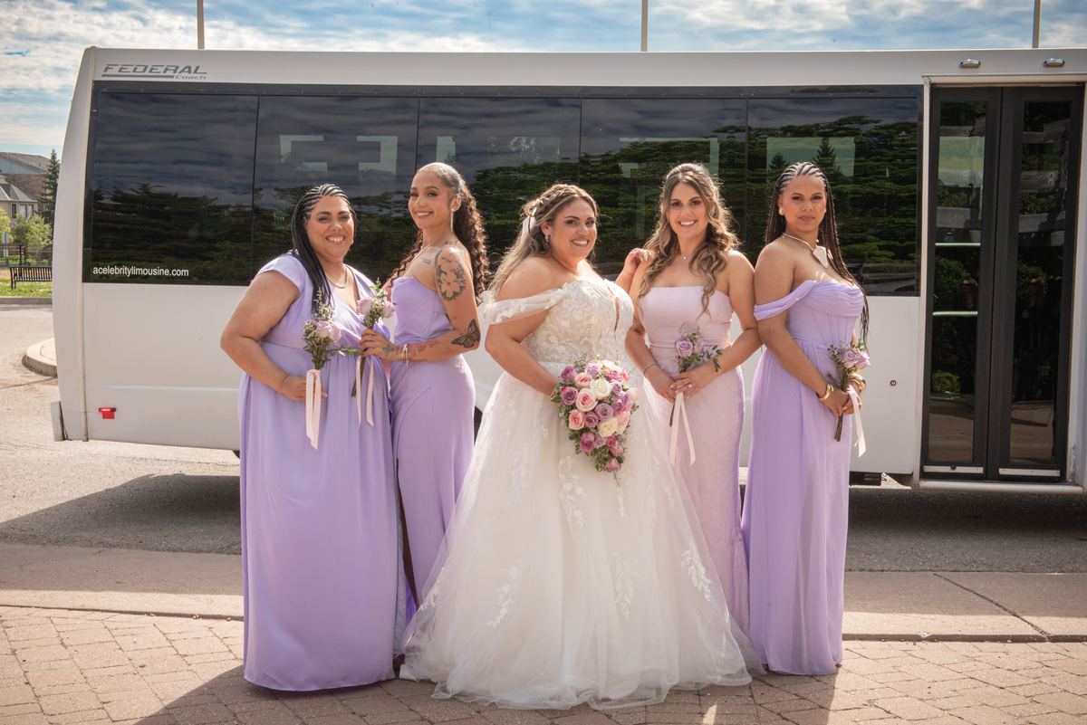 A bride in white with four lavender-clad bridesmaids poses joyfully in front of a tour bus on a sunny day, holding bouquets.