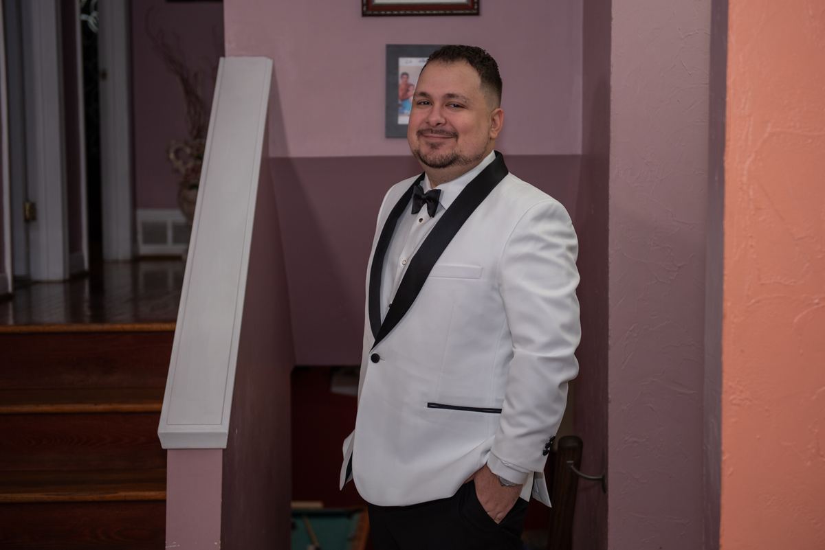 A man in a white tuxedo stands confidently, smiling at the camera with one hand in his pocket near a wooden staircase.