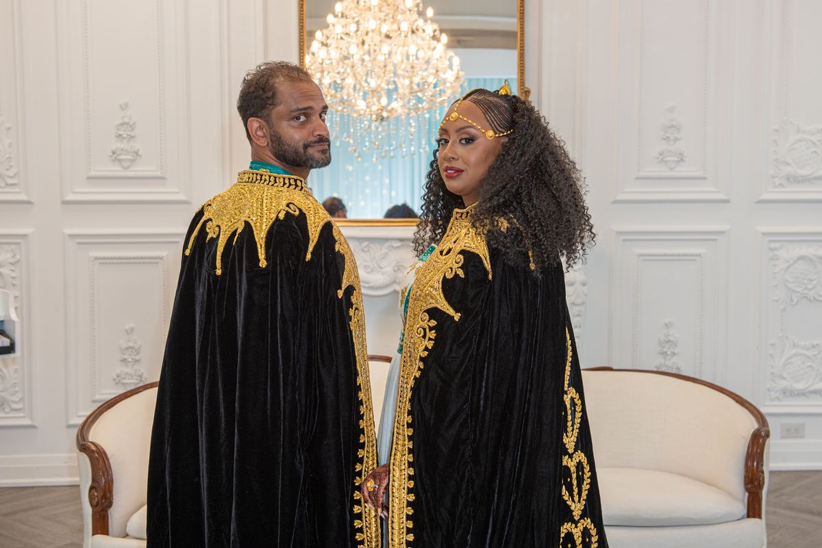 A man and woman in matching black velvet robes with gold embroidery stand facing each other in an ornate room.