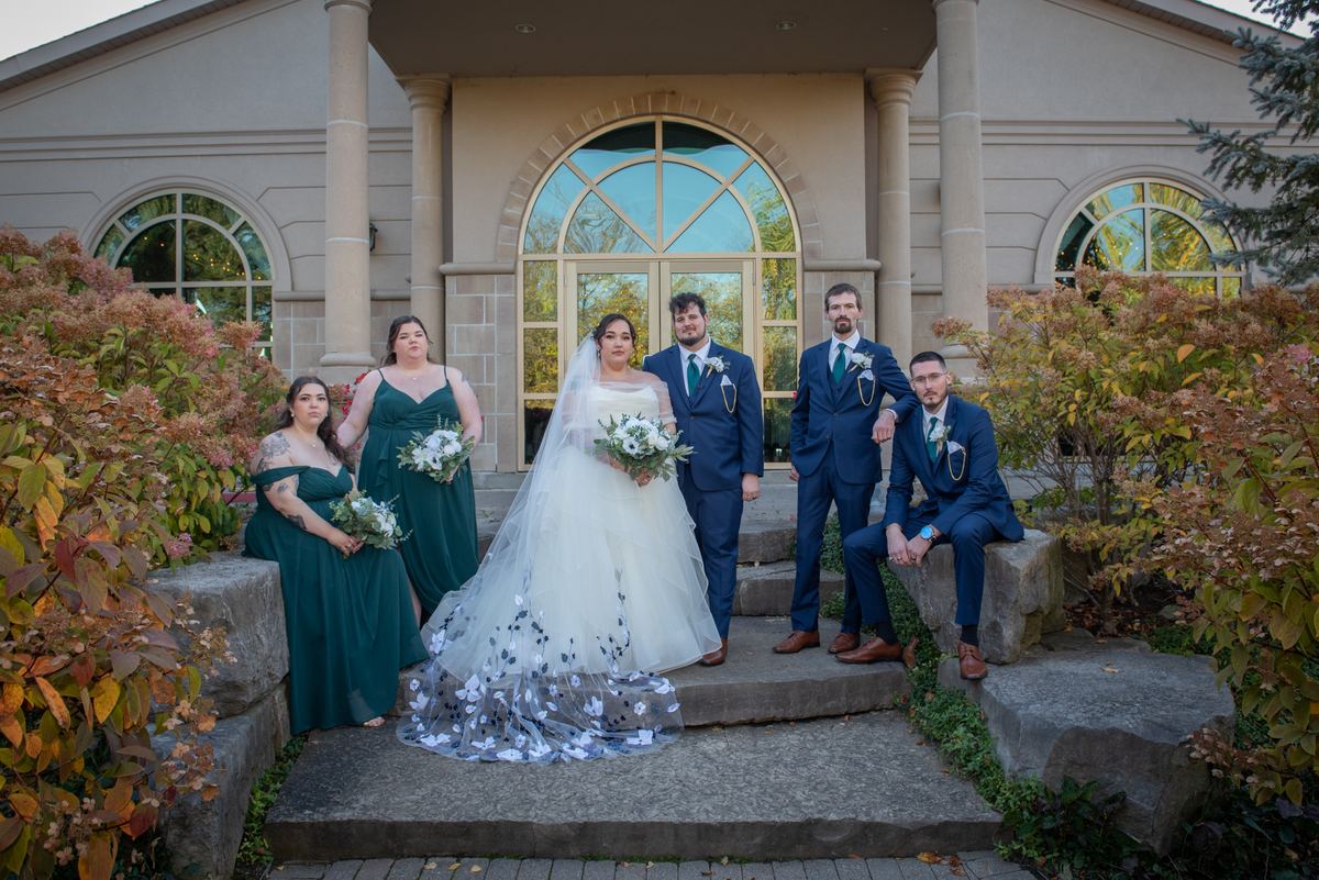A bride and wedding party pose on stone steps outside a stately building, bathed in late afternoon light.