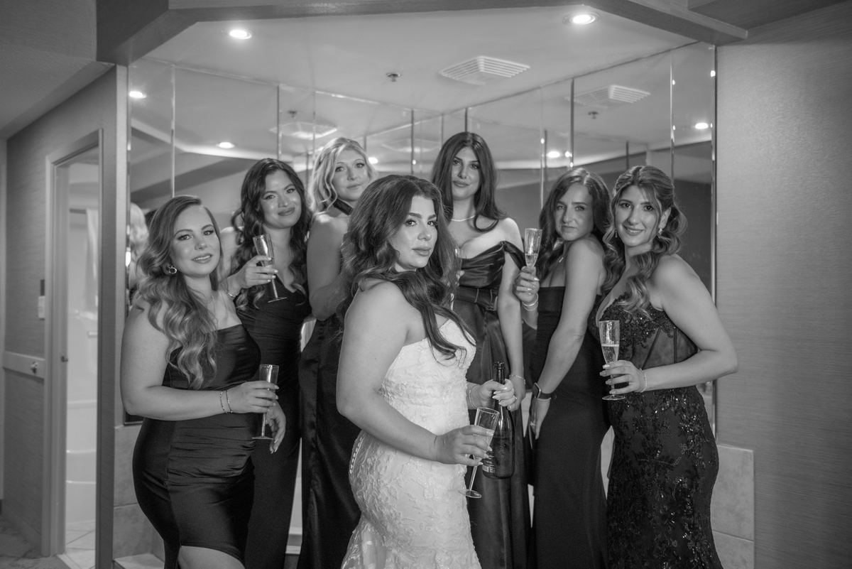 A bride and her bridesmaids stand with champagne flutes in an elegantly lit room with mirrored walls.