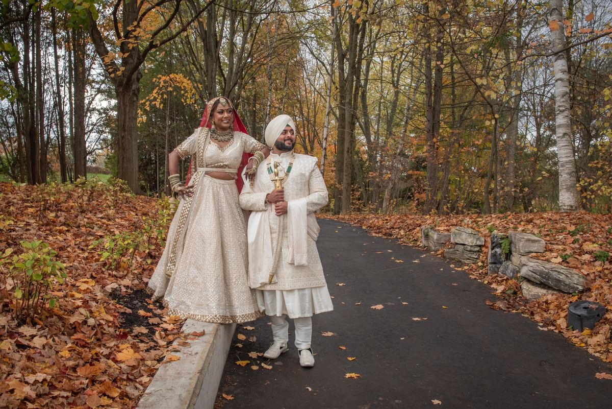 A newlywed couple in traditional South Asian attire stands on a forest path, smiling amid autumn leaves and diffused light.