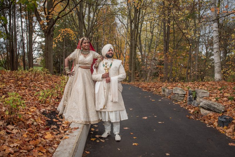 A newlywed couple in traditional South Asian attire stands on a forest path, smiling amid autumn leaves and diffused light.