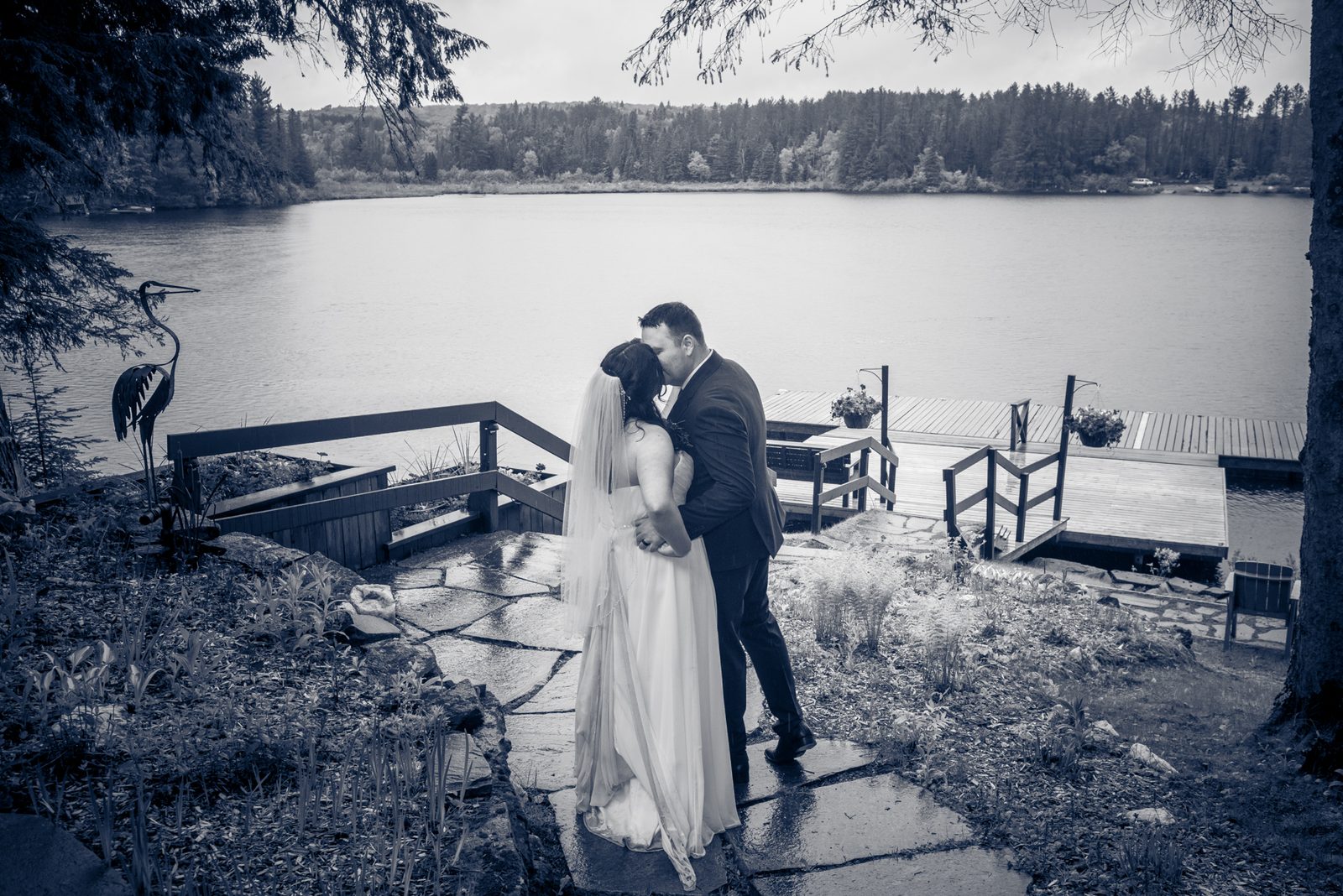 Elise and Alex share an intimate kiss on a stone path beside a wooden dock overlooking a calm lake, framed by trees.